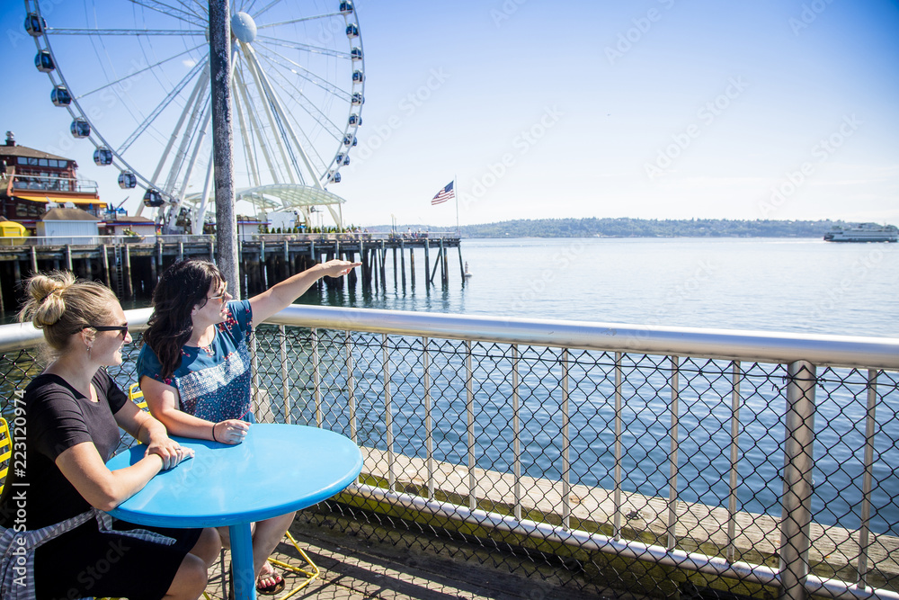 Two women looking out at the Seattle harbor on a sunny day. Sitting at a table on the waterfront near the Ferris Wheel and pier - obrazy, fototapety, plakaty 