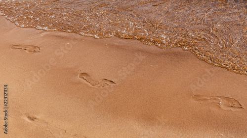 Footprints of bare feet in the sand along a river bank with an oncoming wave