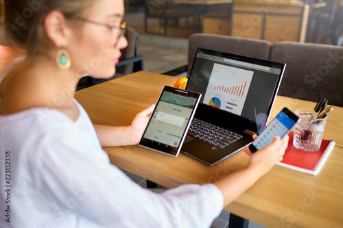 Mixed race woman in glasses working with multiple electronic internet devices. Freelancer businesswoman has tablet and cellphone in hands and laptop on table with charts on screen. Multitasking theme photo