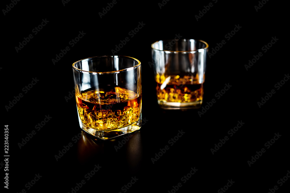 Two glasses of whiskey or whisky with ice on black background.