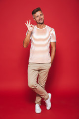 Full length photo of brunette man in striped t-shirt smiling and showing ok sign, isolated over red background