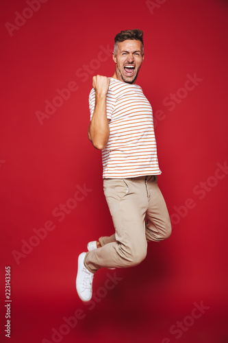 Full length photo of successful man in striped t-shirt screaming and clenching fists, isolated over red background