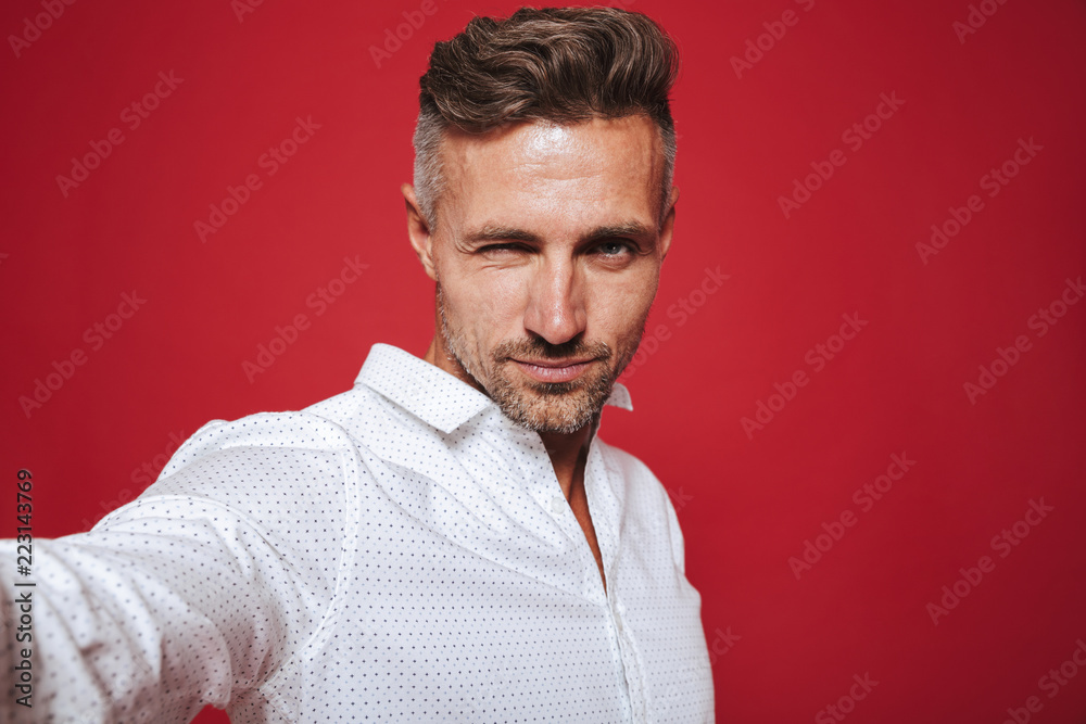 Obraz premium Attractive bearded man 30s in white shirt taking selfie photo, isolated over red background