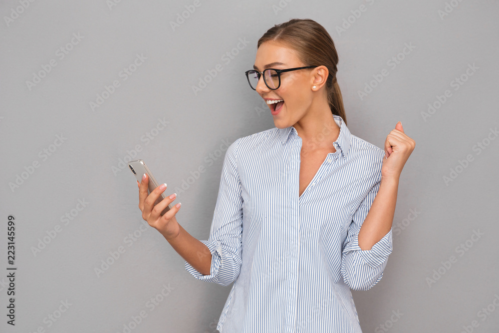 Beautiful happy business woman standing over grey wall background using mobile phone make winner gesture. Beautiful happy business woman standing over grey wall background using mobile phone make winner gesture.