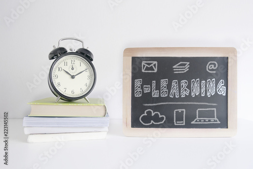 Alarm clock on a pile of books next to a blackboard with the text E-LEARNING written on it. Empty copy space for Editor's text.