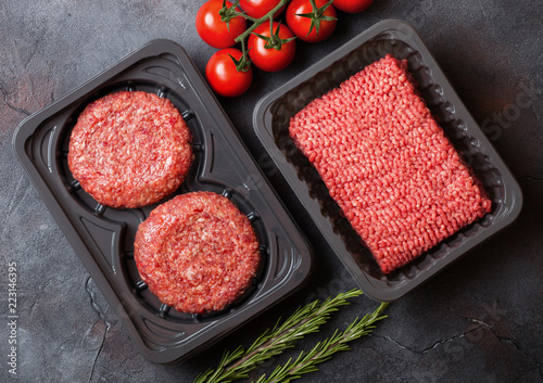 Plastic tray with raw minced homemade meat beef burgers with spices and herbs. Top view and space for text on stone kitchen table background with tomatoes