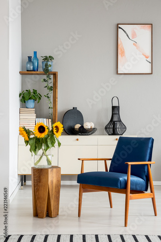 Blue armchair next to wooden table with sunflowers in grey flat interior with poster. Real photo