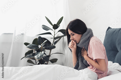 Fototapeta sick young woman sitting in bed and having cough while suffering from sore throa