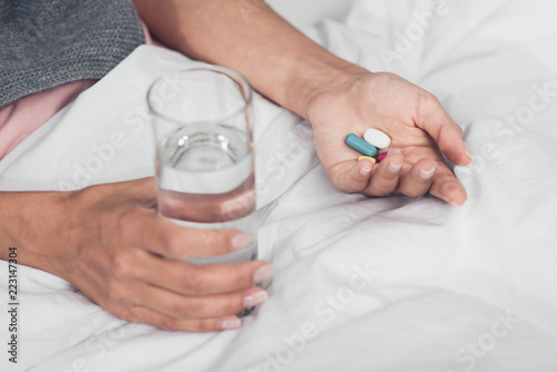 cropped shot of sick woman holding pills and glass of water while lying in bed