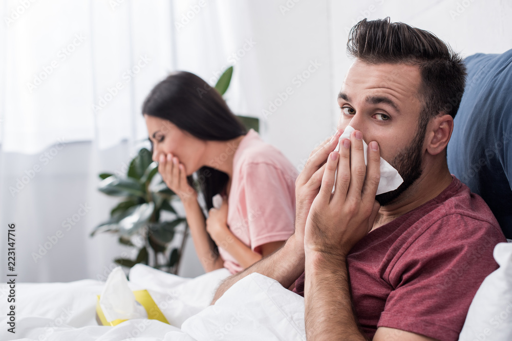diseased young sick couple sneezing into paper napkins while sitting in bed diseased young sick couple sneezing into paper napkins while sitting in bed