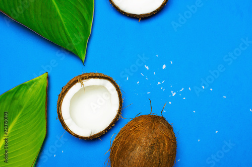 Ripe coconut and tropical leaves on blue colored background, minimal flat lay style top view. Pop art design, creative summer and food concept. Tropical fruit whole and half abstract background