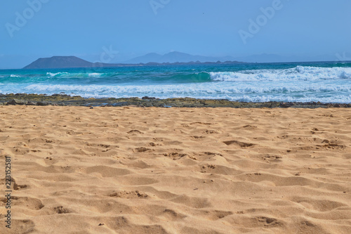 Landscape of golden sand and stones on the shore with the sea and waves and mountains in the background on a clear day in Corralejo, Fuerteventura, Canary Islands, Spain