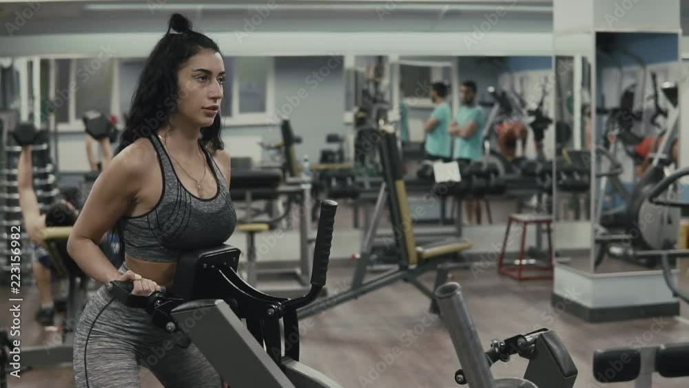 Fitness girl doing exercise in block simulator in slow motion. Brunette girl in gray sportswear in gym with copy space Fitness girl doing exercise in block simulator in slow motion. Brunette girl in gray sportswear in gym with copy space