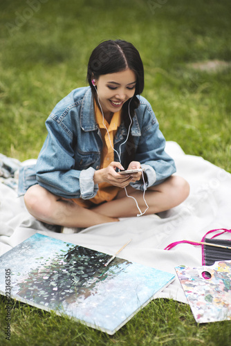 Hipster girl in casual wear and eyeglasses checking new feeds in social networks on break in park, young woman sending text message using cellular and 4G intenret sitting on grass during free time photo