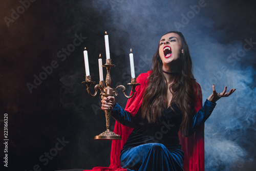 Wall Mural woman in red cloak holding vintage candelabrum and showing vampire fangs on dark