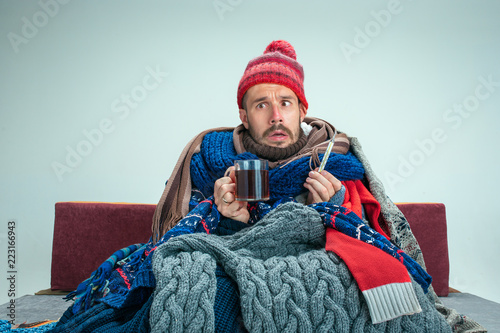 Bearded sick man with flue sitting on sofa at home or studio with cup of tea covered with knitted warm clothes. Illness, influenza concept. Relaxation at Home. Healthcare Concepts.