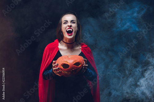 mystic woman in vampire costume holding jack o lantern on dark background with smoke