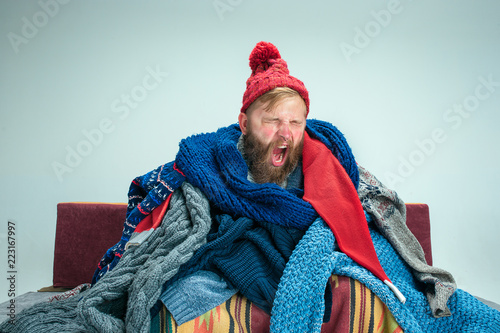 Bearded sick man with flue sitting on sofa at home or studio covered with knitted warm clothes. Illness, influenza concept. Relaxation at Home. Healthcare Concepts.