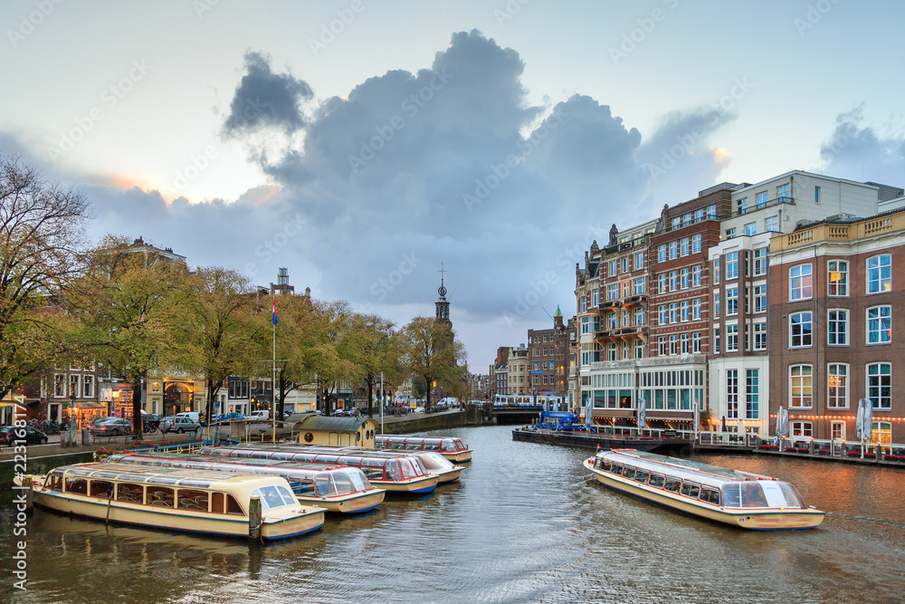Beautiful cityscape view towards the Mint tower (Munttoren) with tourist canal boats at the Amstel river in Amsterdam, the Netherlands, with ominous clouds around sunset