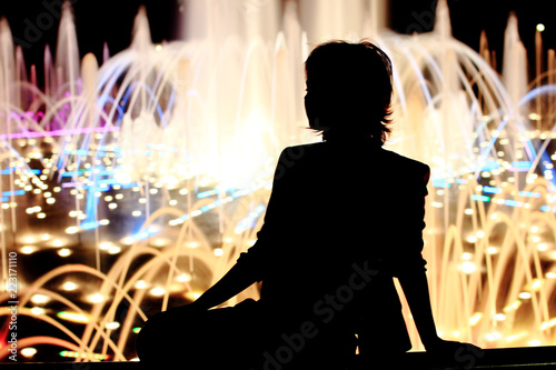 Fine girl on the street. Portrait, full length, sitting.Yonge woman on pastel background with jet fountain. Black silhouette, text space.