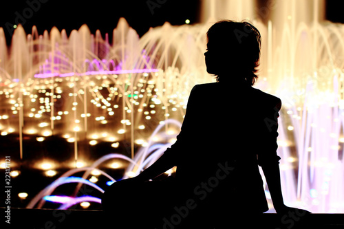 Fine girl on the street. Portrait, full length, sitting.Yonge woman on pastel background with jet fountain. Black silhouette, text space.