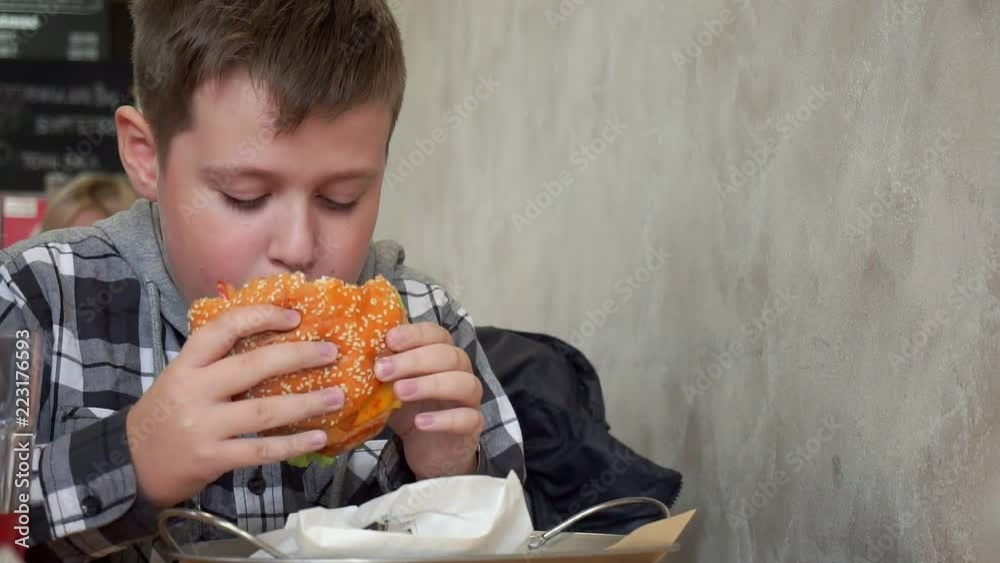 A teenager in a cafe eating a hamburger. He is hungry, tasty and juicy hamburger A teenager in a cafe eating a hamburger. He is hungry, tasty and juicy hamburger
