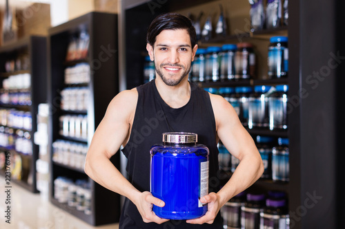 Young bodybuilder showing power and holding pot  of sport nutrition products in shop