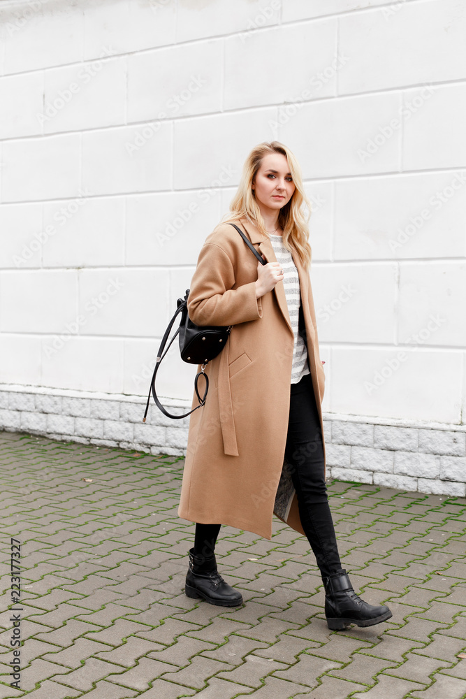 Beautiful young stylish blonde woman wearing long beige coat, black boots  and backpack walking through the city streets. Trendy casual outfit. Street  fashion. Stock Photo | Adobe Stock