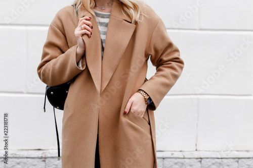 Beautiful young stylish blonde woman wearing beige coat and black backpack posing near white street wall. Trendy casual outfit. Street fashion. Details of everyday look. photo