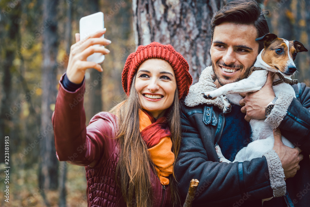 Naklejka premium Woman and man with their dog on autumn walk taking a phone selfie posting it online on social media