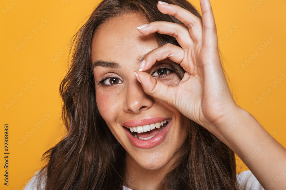 Portrait of happy woman 20s with long hair laughing and looking through hole made with her fingers, isolated over yellow background Portrait of happy woman 20s with long hair laughing and looking through hole made with her fingers, isolated over yellow background