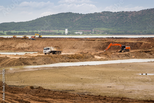 Excavator and truck in the area of construction with mountrain, river and sky background.