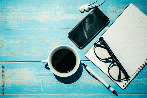 Cup of black coffee and smart phone with office supplies; pen, notebook and eyes glasses on blue wooden table background. Top view with copy space