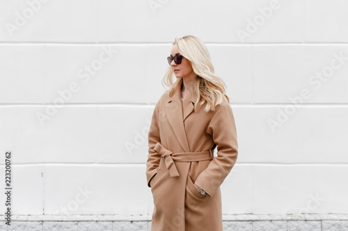 Beautiful young stylish blonde woman wearing beige coat and black sunglasses standing near white street wall. Trendy casual outfit. Street fashion. photo