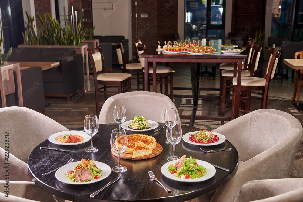 A solemn table with glasses and a variety of different dishes from the chief chef in the foreground. Buffet table with appetizers, canapes and sandwiches in the background.