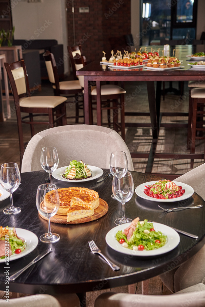 A solemn table with glasses and a variety of different dishes from the chief chef in the foreground. Buffet table with appetizers, canapes and sandwiches in the background.