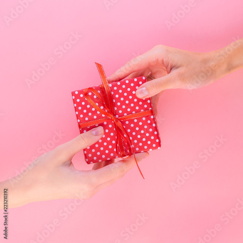 Two women holding a gift in a red gift box with a bow, passing it from one to the other, pink background, top view