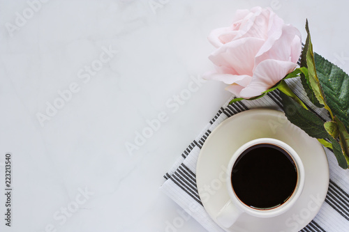 Cup of black coffee with saucer and sweet pink rose on napery against white marble background for drinks and beverage concept