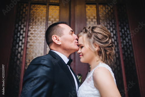 Smiling and beautiful newlyweds look at each other and kiss, holding hands, near the ancient door in a chic interior. A wedding portrait of an angry groom and a young bride. Wedding.