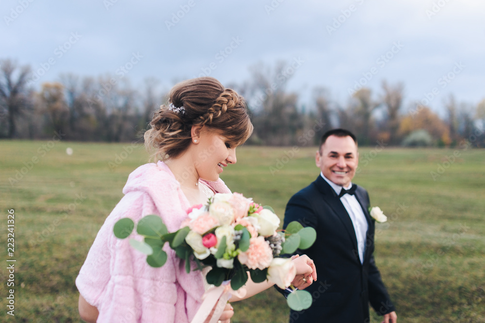 An adult groom and pretty bride strolling along the green field against the backdrop of a pine forest and smiling. Wedding portrait close-up of beautiful newlyweds. An adult groom and pretty bride strolling along the green field against the backdrop of a pine forest and smiling. Wedding portrait close-up of beautiful newlyweds.