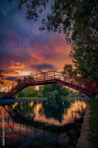 Fototapeta Naklejka Na Ścianę i Meble -  Dramatic sunset sky with vibrant colors in a park with a red bridge over a lake