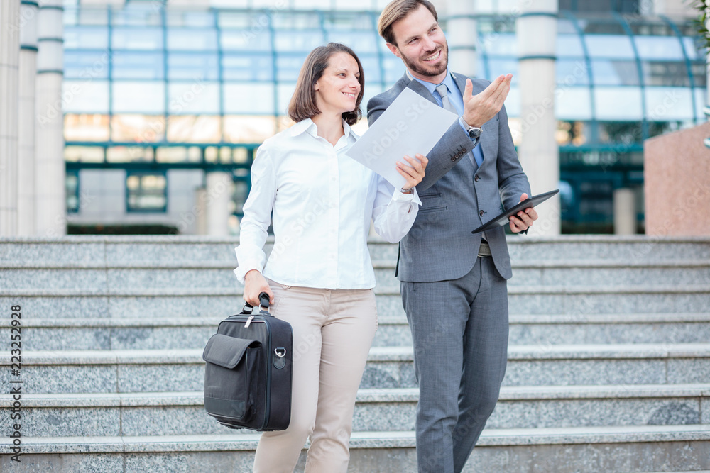 Successful young male and female businesspeople walking down the stairs in front of an office building. Woman is carrying briefcase and paper documents and man is holding a tablet in his hand Successful young male and female businesspeople walking down the stairs in front of an office building. Woman is carrying briefcase and paper documents and man is holding a tablet in his hand