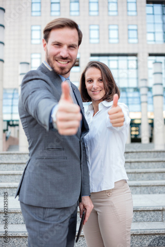 Young male and female businesspeople standing in front of an office building and giving thumbs up. Focus on a woman © Ivan