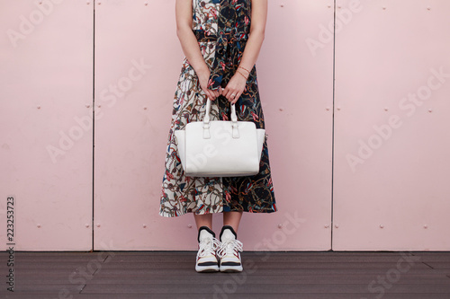 Woman in a fashionable dress with a white stylish bag stand near the pink wall. Women's Fashion Sneakers
