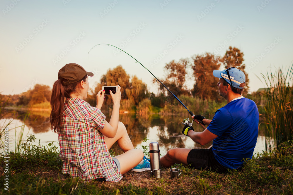 Young couple fishing and drinking tea on river at sunset. Woman