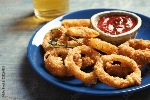 Plate with homemade crunchy fried onion rings and tomato sauce on wooden background, closeup
