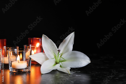 White lily and burning candles on table in darkness, space for text. Funeral symbol
