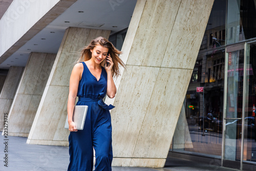 Young Eastern European American Woman talking on cell phone, traveling, working in New York City, wearing blue sleeveless jumpsuit, carrying laptop computer, walking on street outside office building photo