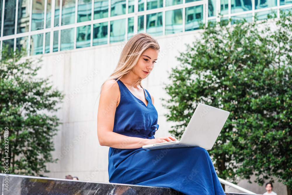 Way to Success. Young Beautiful Eastern European American Woman studying in New York, wearing blue sleeveless jumpsuit, sitting outside on campus, looking down, reading, working on laptop computer..