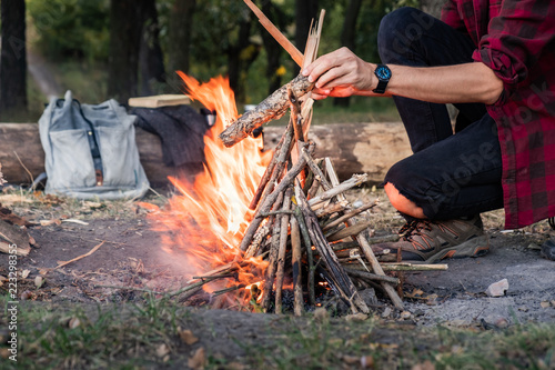 Making campfire at a forest. Going into the wild concept: camping place with vintage backpack, thermos and male in casual clothes puts pieces of wood into fire.